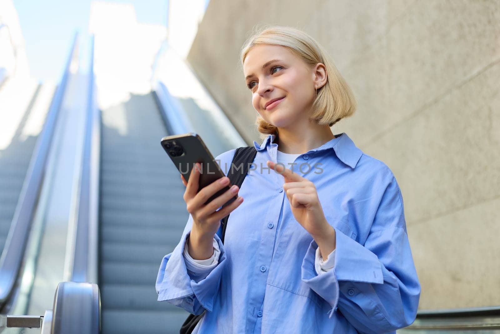 Close up portrait of young blond woman in city centre, standing near escalator, checking her mobile phone, reading message on smartphone by Benzoix