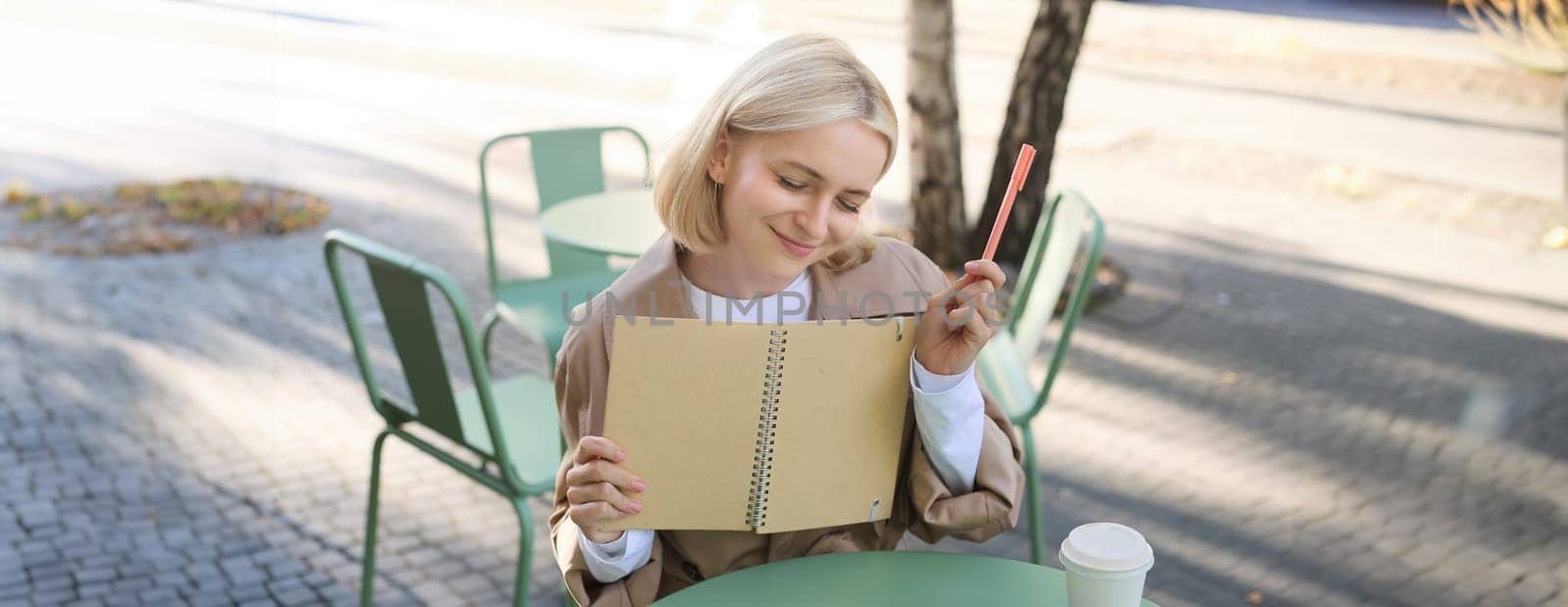 Portrait of young creative woman, doing sketches outdoors, sitting in coffee shop with notebook and pen, drawing art and smiling.