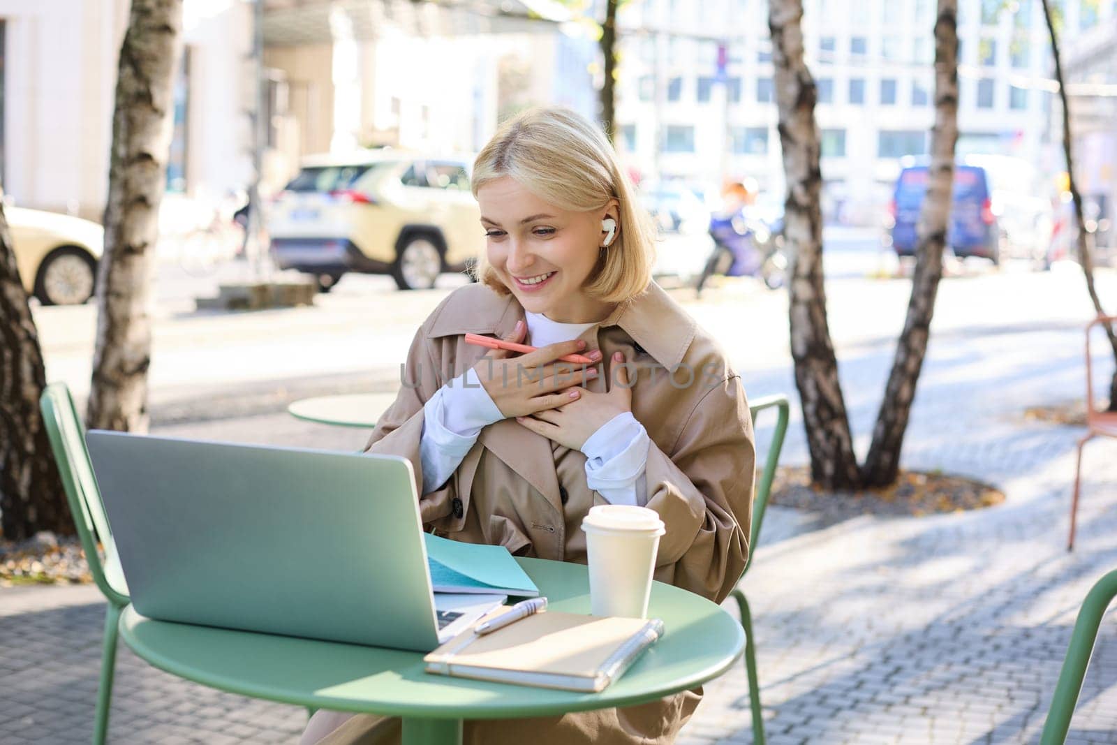 Portrait of young happy woman, looking pleased and surprised at laptop screen, sitting in outdoor cafe, street coffee shop, holding hands on chest and smiling grateful.