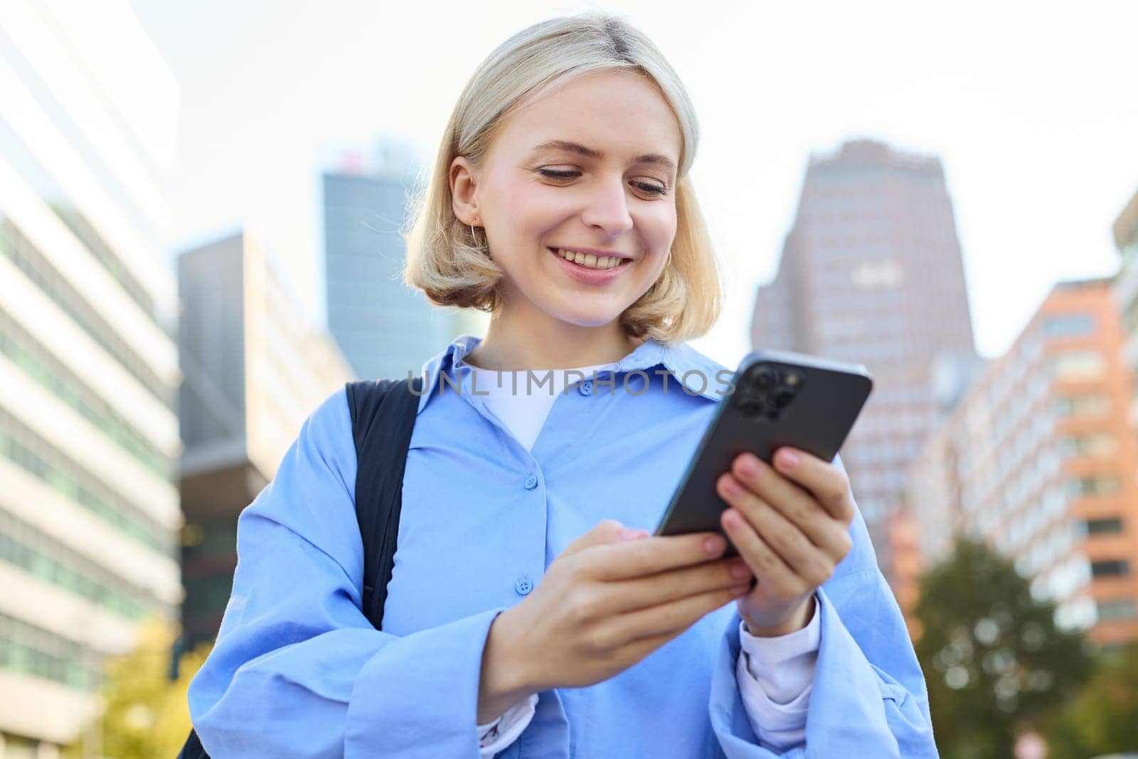 Close up portrait of stylish young blonde woman, standing on street, checking her mobile phone, using smartphone app to get around town, looking at online map for directions.