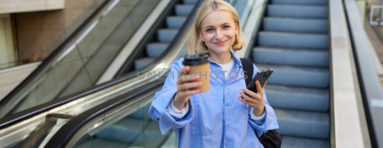 Portrait of smiling, beautiful young woman with smartphone, standing on bottom of escalator, giving you cup of coffee, brought takeaway by Benzoix