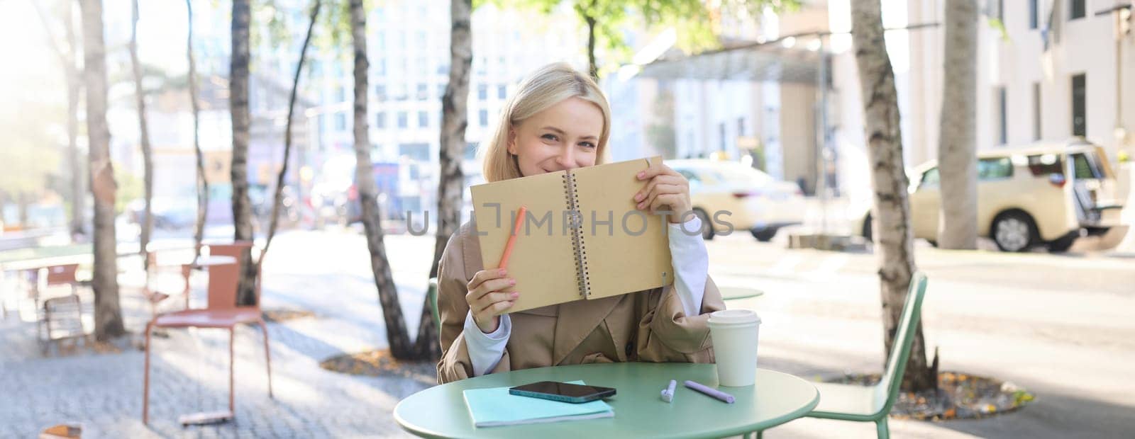 Image of young beautiful woman, student doing homework in an outdoor coffee shop, holding journal or planner, writing in notebook, smiling happily by Benzoix