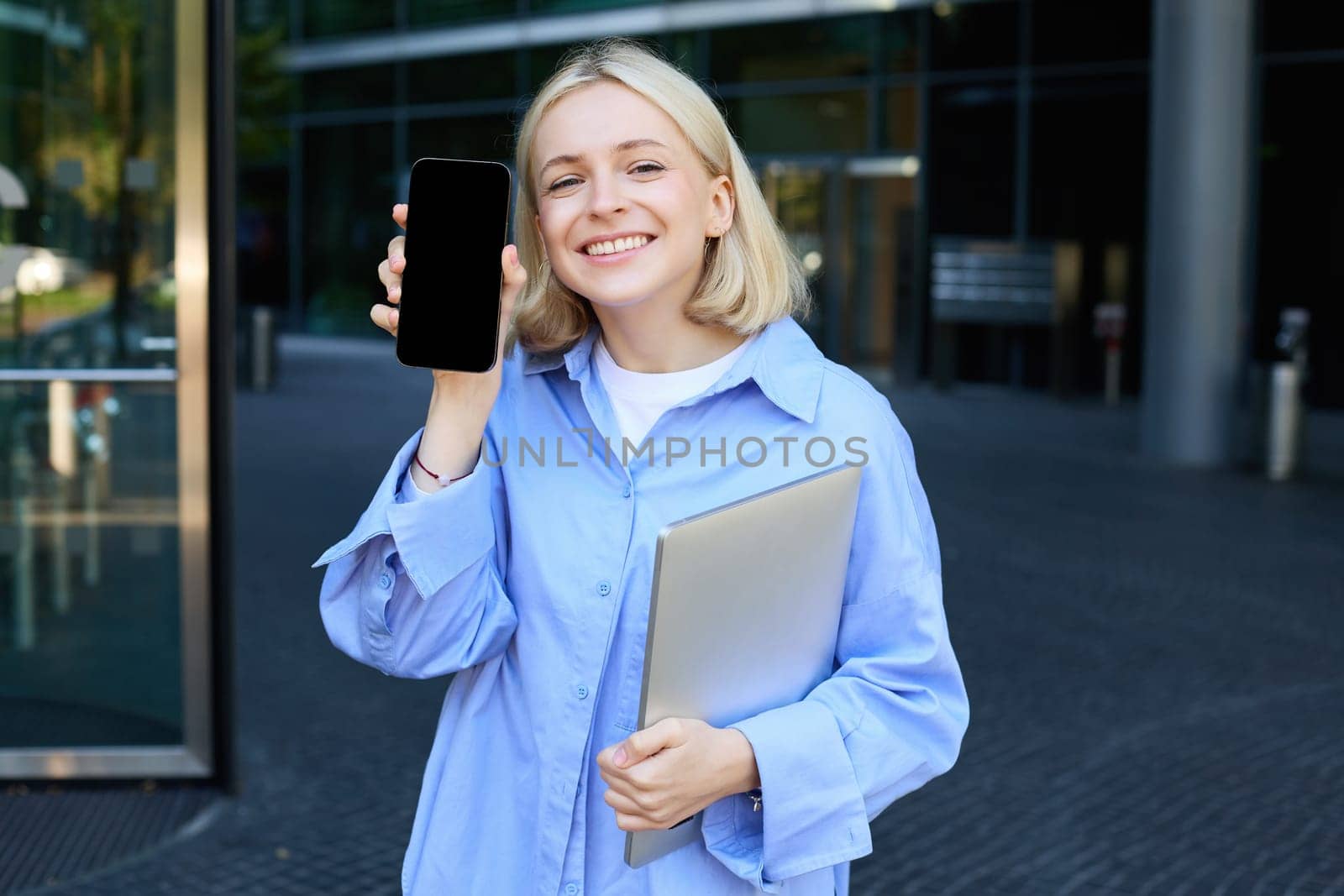 Happy smiling woman showing smartphone screen, demonstrating mobile phone display, standing in college outdoors, posing near campus with laptop and telephone by Benzoix