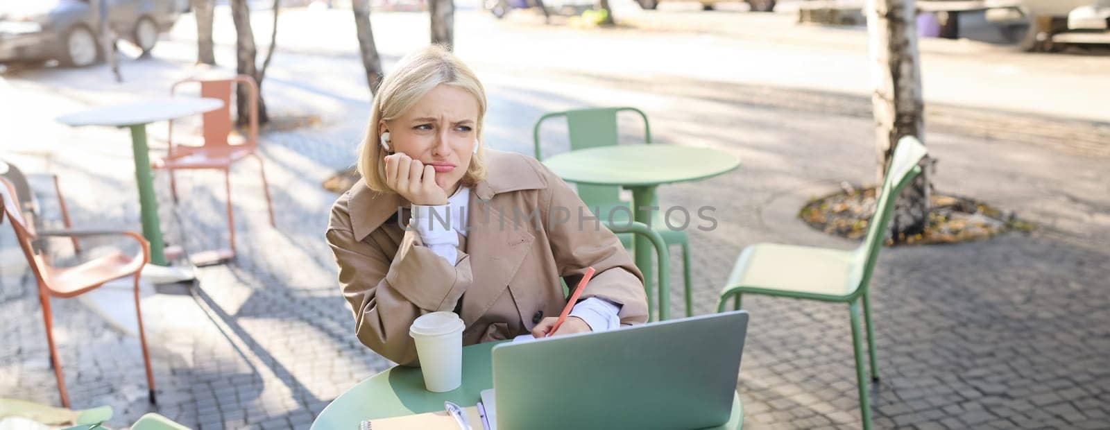 Portrait of young blond woman with laptop, sitting on street, drinking coffee in outdoor cafe, working, wearing wireless headphones, doing homework, freelancing by Benzoix