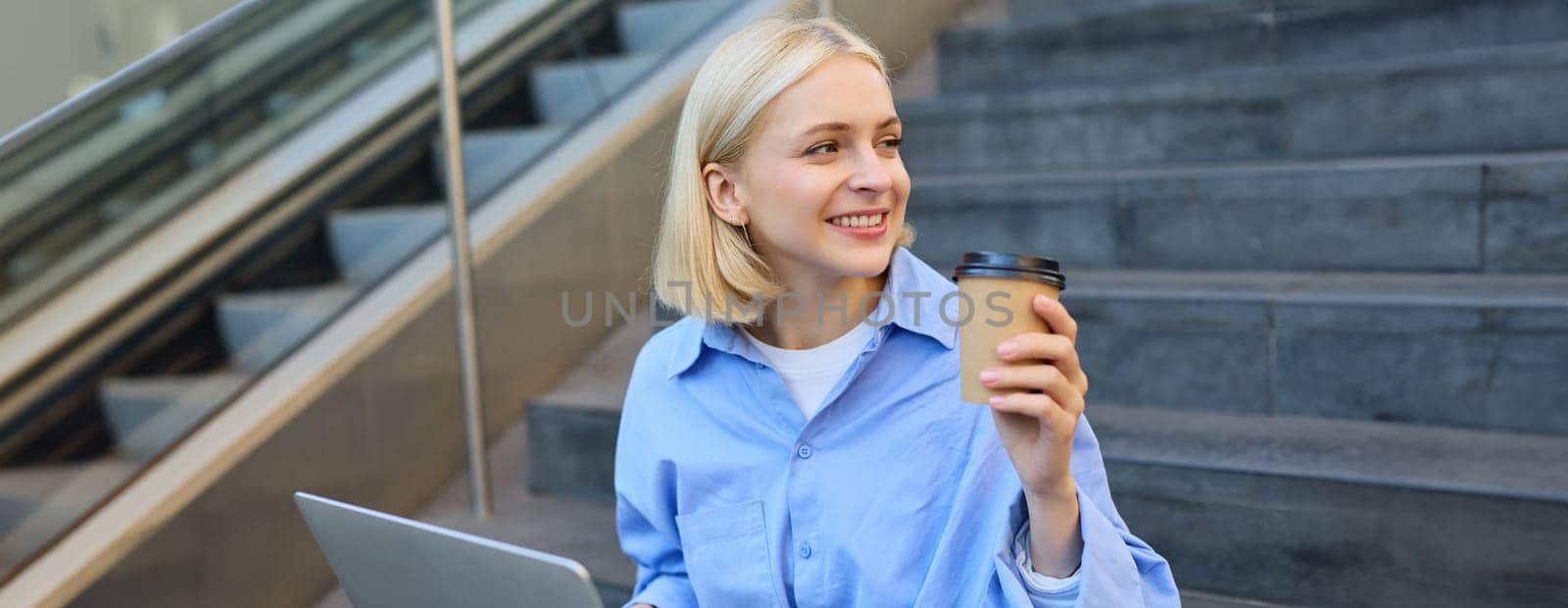 Portrait of blond smiling woman, drinking coffee, using laptop, sitting on campus stairs outside of building, studying, e-learning, connecting to wifi.