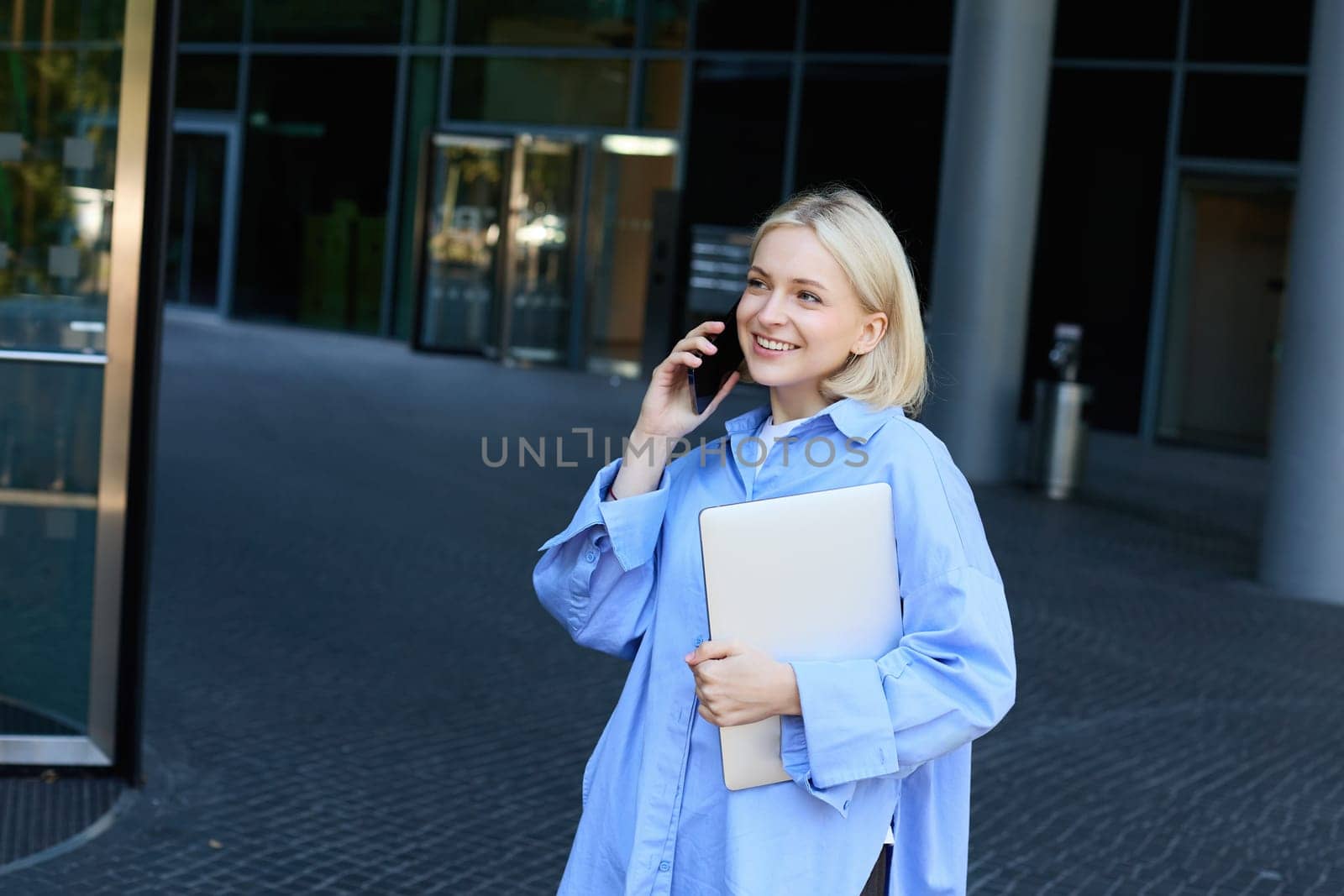 Image of young office woman, employee standing near company building with documents and notebooks, answers phone call, talking on smartphone and smiling, wearing blue collar shirt by Benzoix