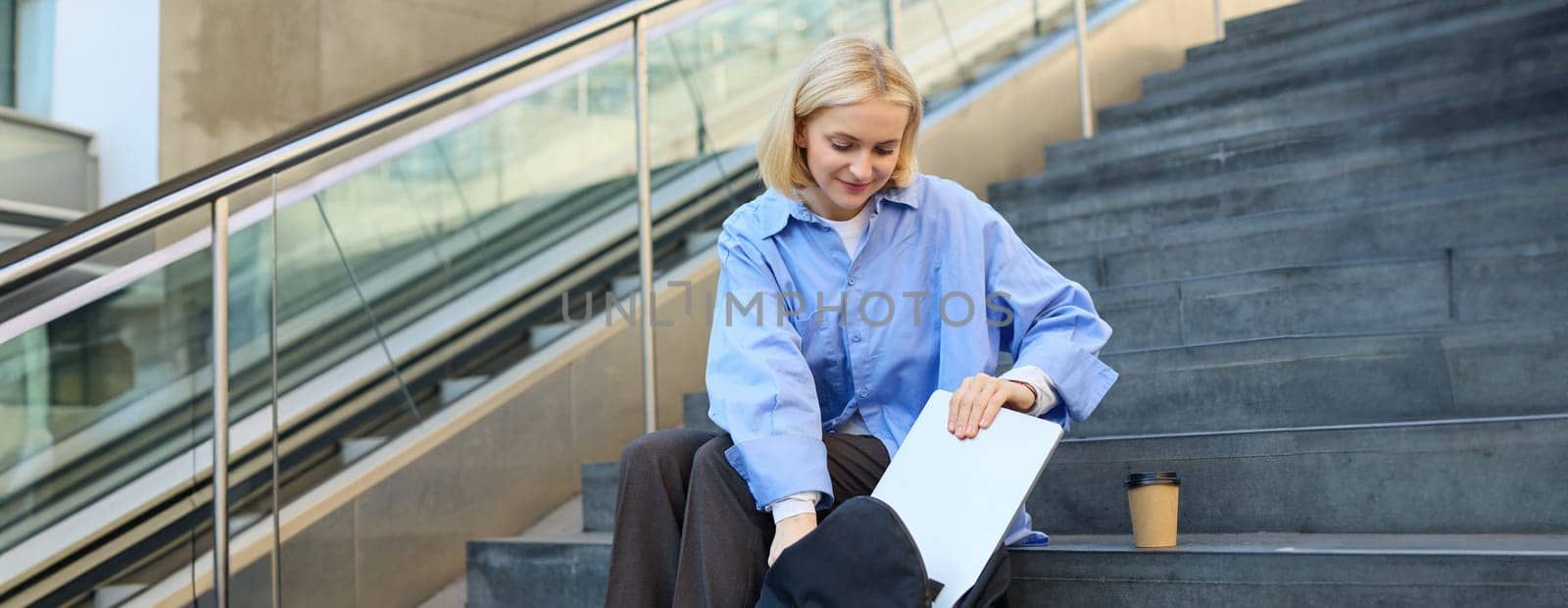 Image of smiling young woman, student packing her laptop in backpack, sitting on public stairs with cup of coffee, hurry to lecture by Benzoix