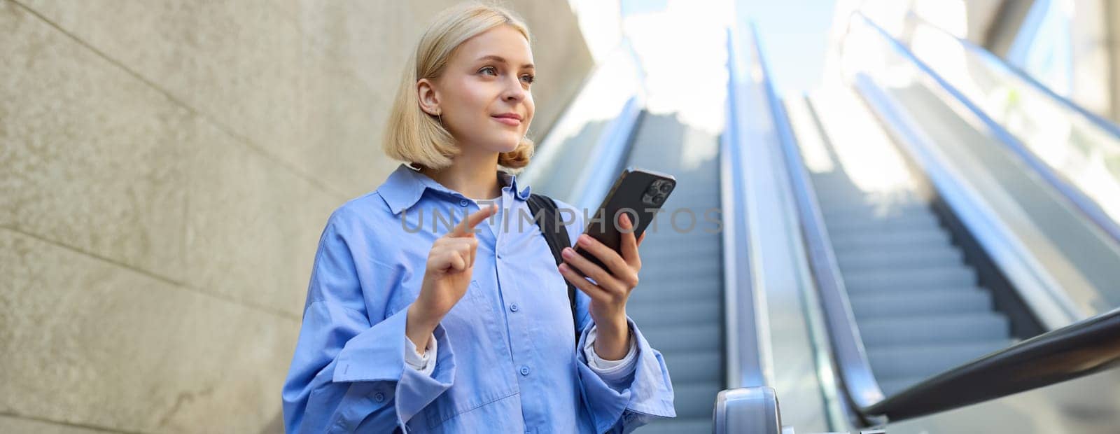 Portrait of beautiful young blond woman in blue shirt, standing near escalator, holding mobile phone, using smartphone, waiting for someone in city centre by Benzoix