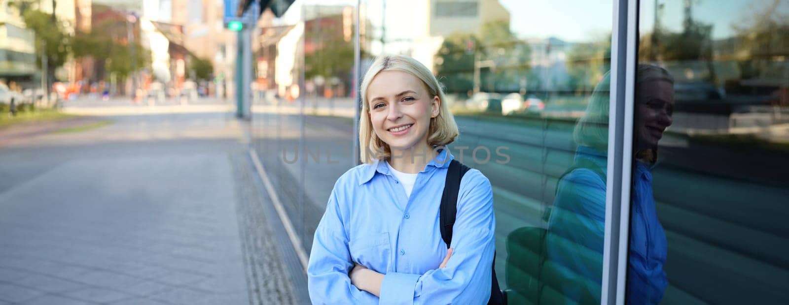 Lifestyle portrait of young smiling woman, student or office employee, standing on street in blue shirt, cross arms on chest and looking confident at camera by Benzoix