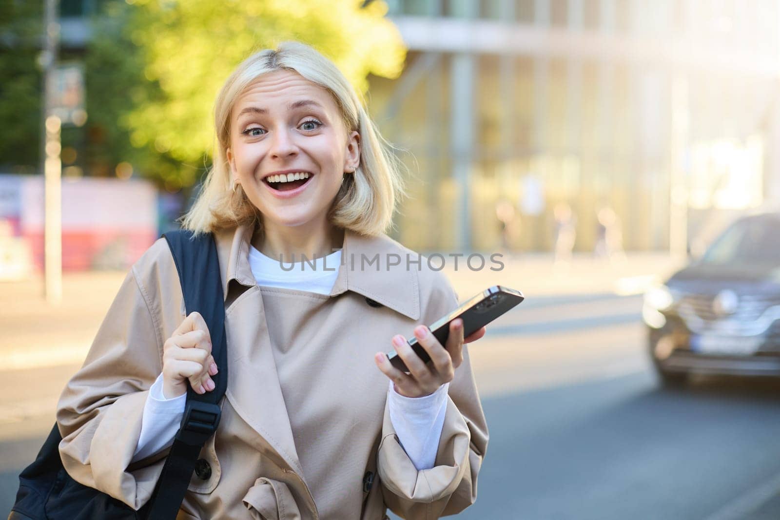 Portrait of excited blonde woman, college student standing on street with mobile phone, holds backpack.