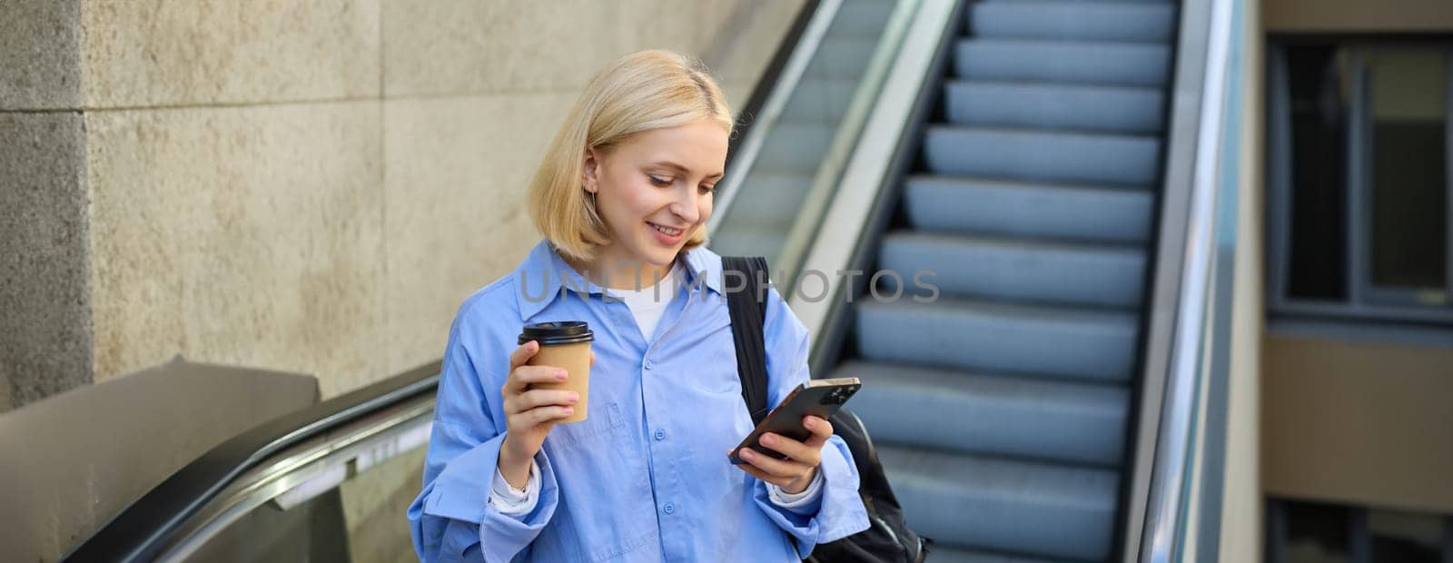 Portrait of blond young woman, reading message on her mobile phone, drinking takeaway coffee, standing near escalator with backpack.