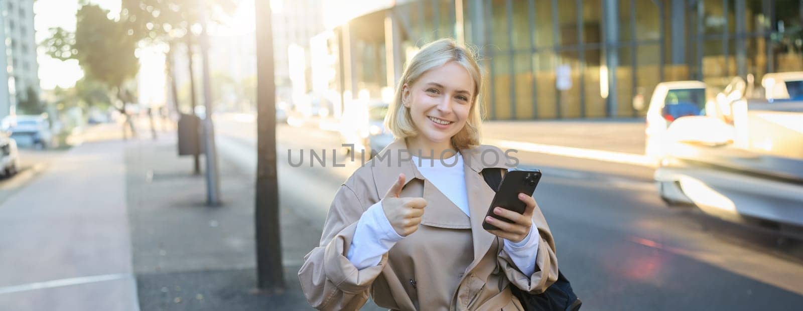Lifestyle portrait of happy smiling woman, girl standing on street with smartphone, showing thumbs up with pleased face, recommending something good.