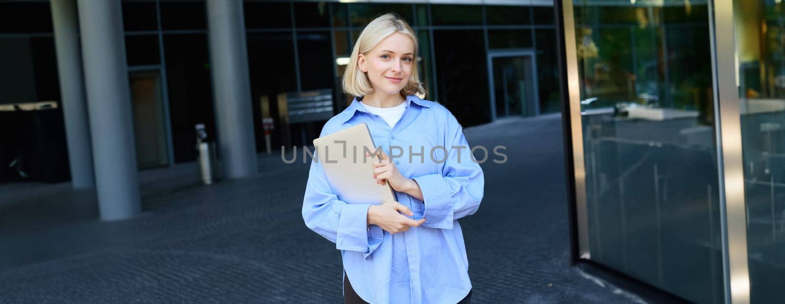 Stylish, modern young woman, student standing on street in campus, heading to lecture with notebook and study material, smiling at camera.