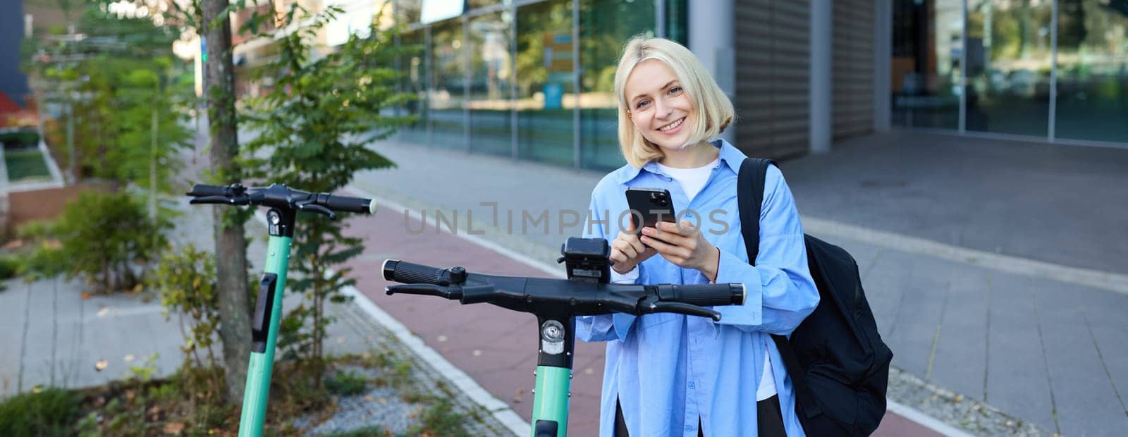 Portrait of smiling young woman unlocks scooter on street, using mobile phone to rent it with smartphone application, riding to university.