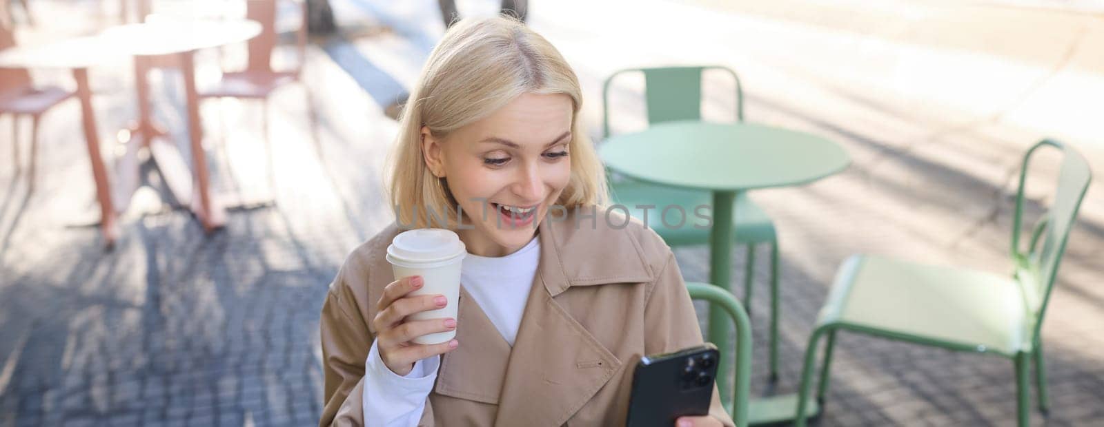 Image of blond female model, looks surprised at her social media account, staring at smartphone with amazed face, drinking coffee outdoors, sitting outside.