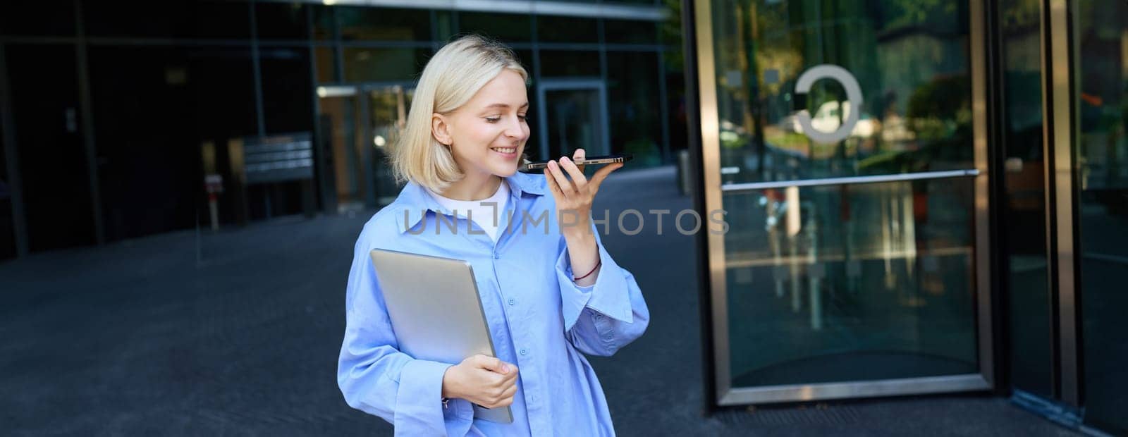 Image of smiling beautiful blond girl, office employee with laptop, records voice message on smartphone app, standing near company building on street.