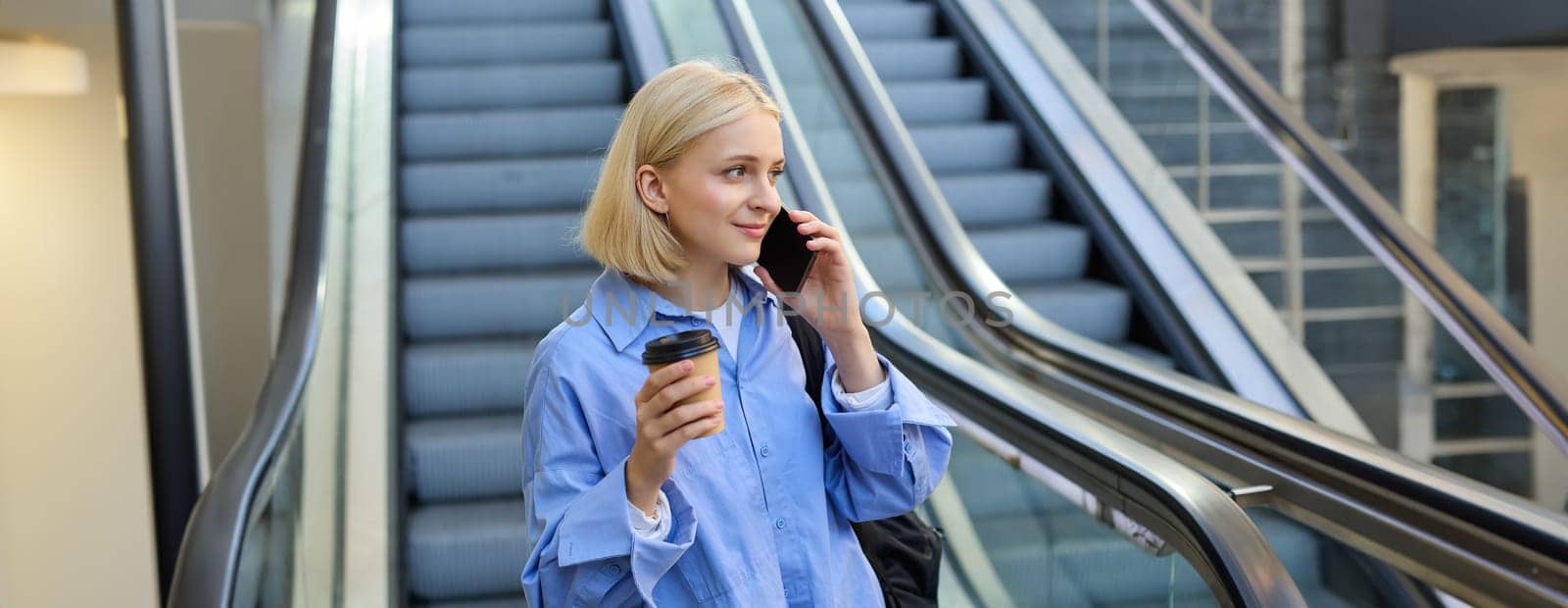 Lifestyle shot of young female student, woman talking on mobile phone, drinking coffee, standing near escalator in city centre.