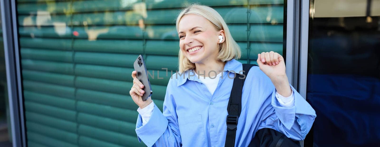 Carefree woman with backpack and mobile phone, wearing wireless earphones, listening to music and smiling, standing on street in city centre, laughing.
