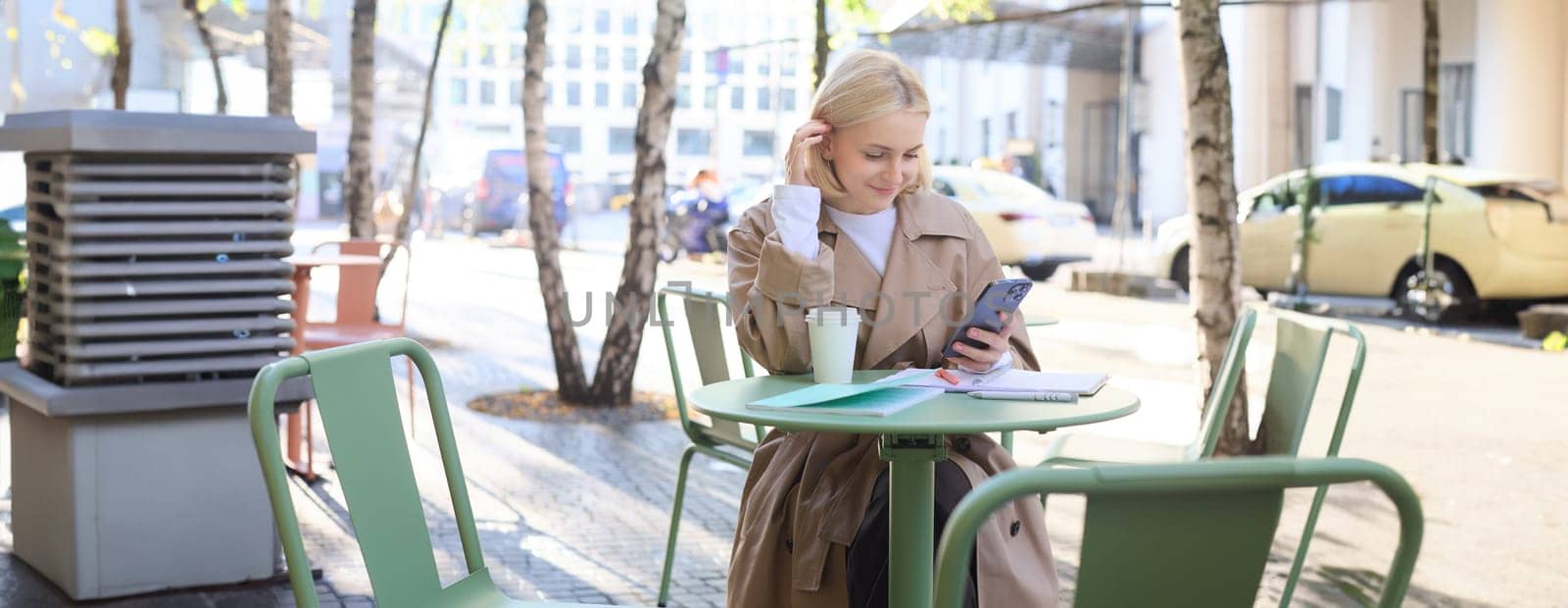 Street style portrait of young smiling woman sitting on street outdoors, holding mobile phone, using smartphone, drinking coffee in local cafe.