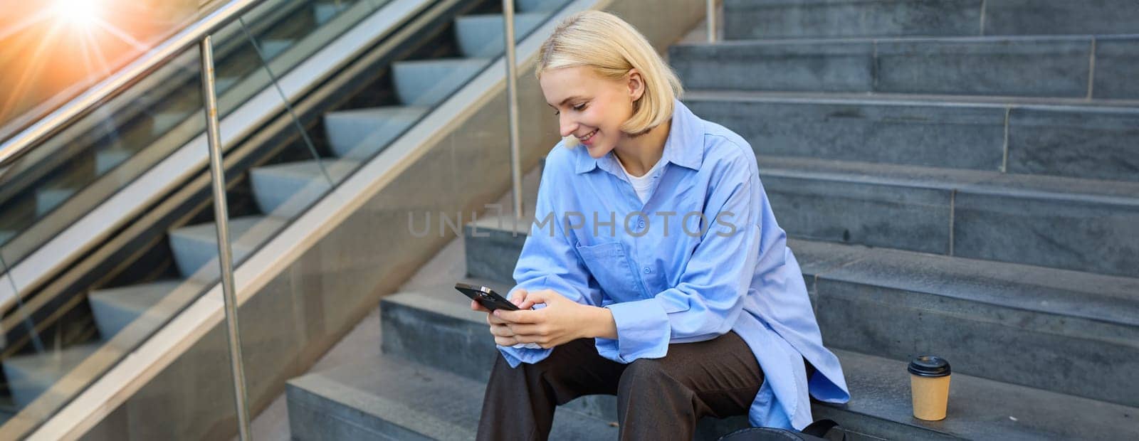Lifestyle portrait of smiling, young woman, sitting on street stairs, looking at mobile phone, reading a message on smartphone, drinking coffee on lunch break.