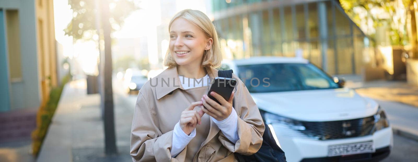 Portrait of young woman using map application on smartphone, student with backpack, walks along street on sunny day, holds mobile phone in hand by Benzoix