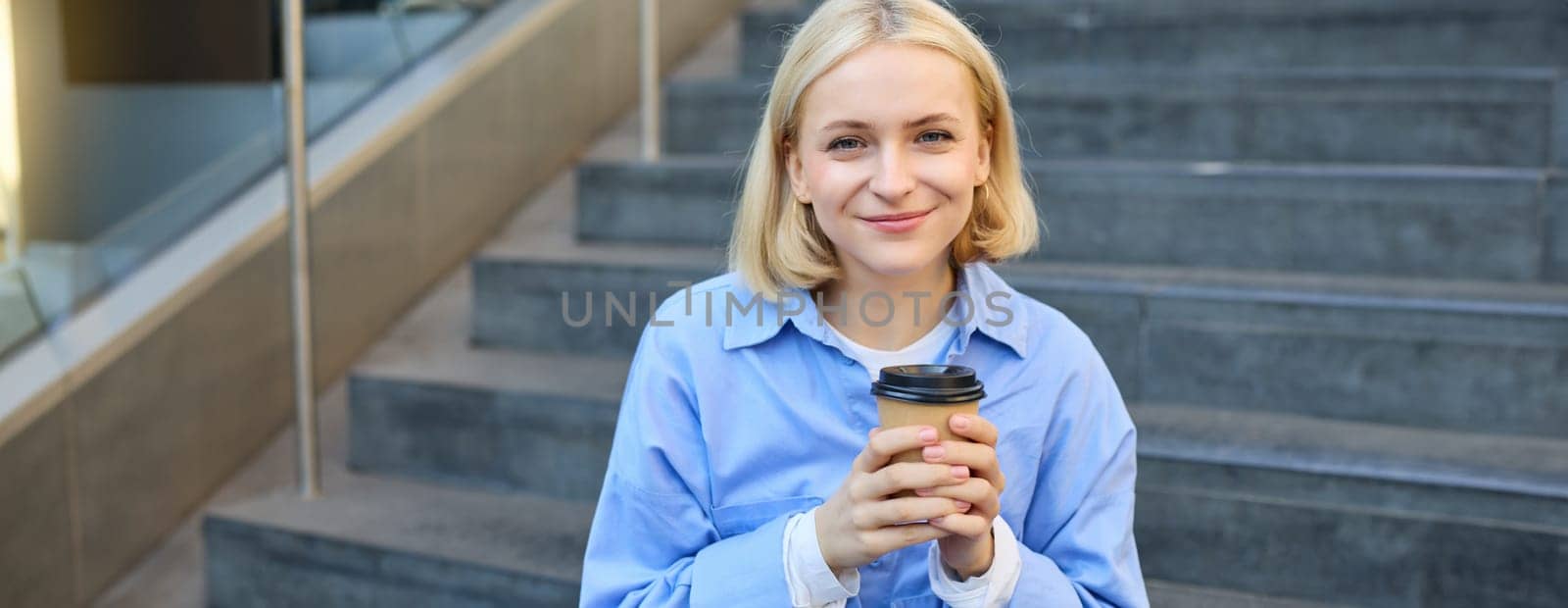 Close up portrait of beautiful, smiling blonde woman, student sitting on stairs outside campus, drinking takeaway coffee, warm-up her hands while holding a cup.