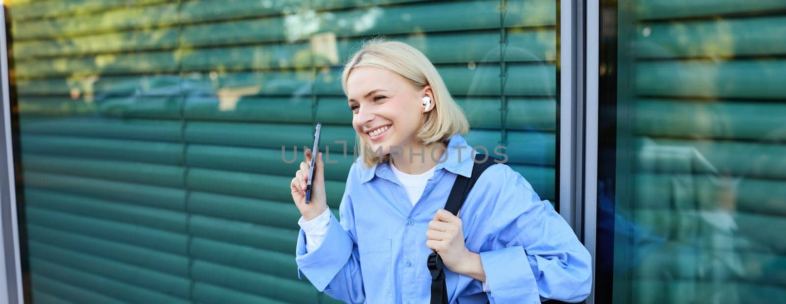 Close up portrait of blond smiling female model, college student in earphones, listening to music on street, holding smartphone, waiting for someone outdoors.