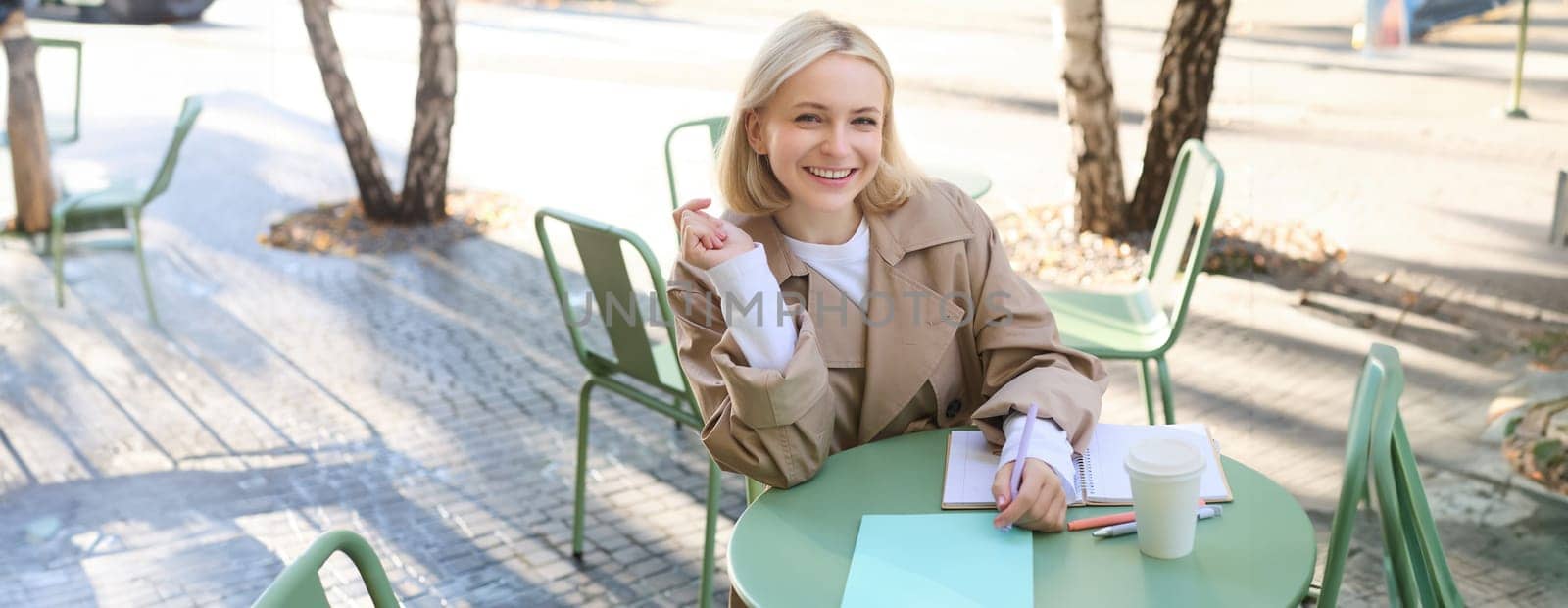 Portrait of carefree, blond cute woman working with documents, drinking takeaway coffee and sitting in cafe outdoors with documents, writing essay, student doing homework.