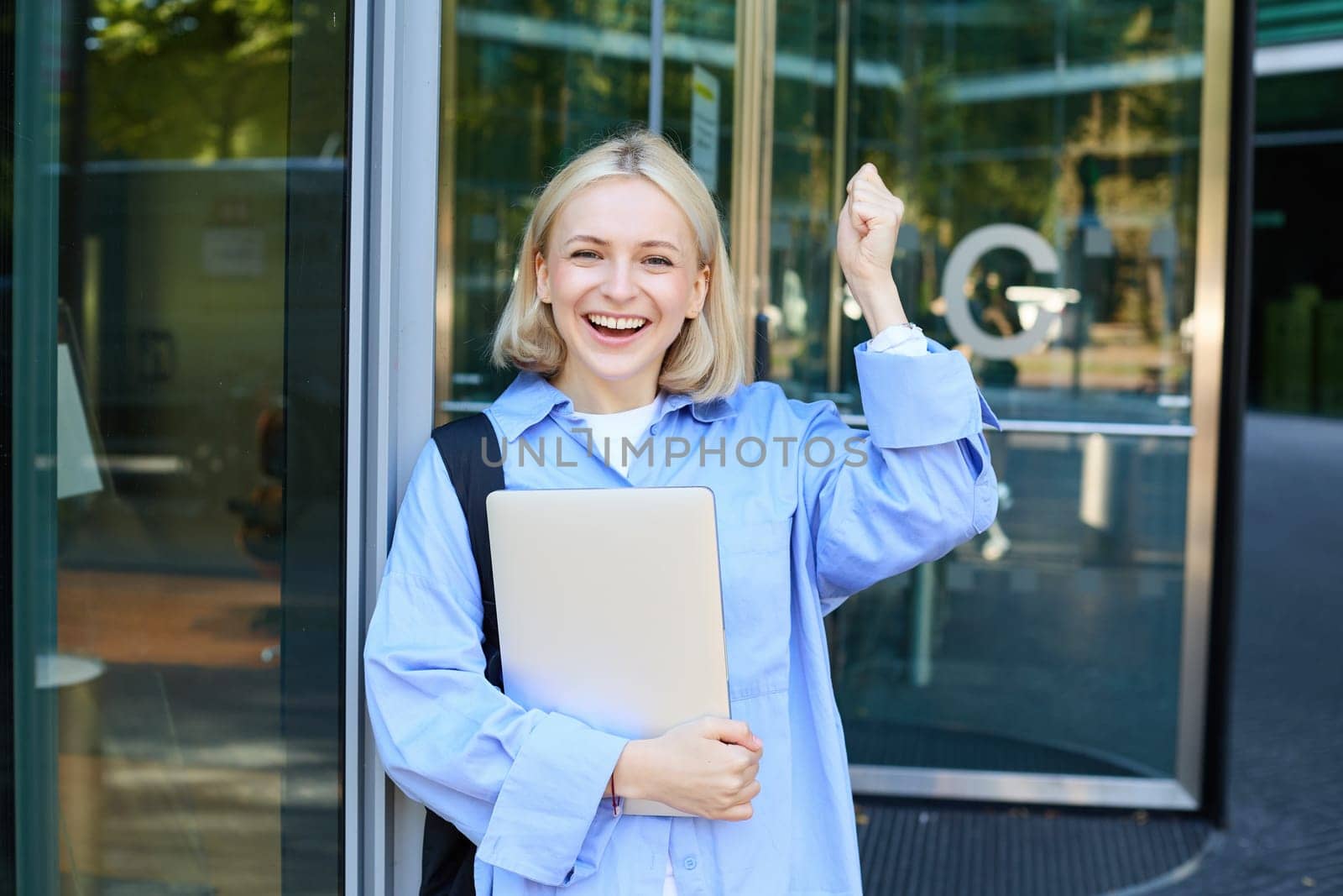 Portrait of happy, enthusiastic blond woman near office building, holding backpack and laptop, raising hands up and cheering, encouraging you, chanting and smiling.