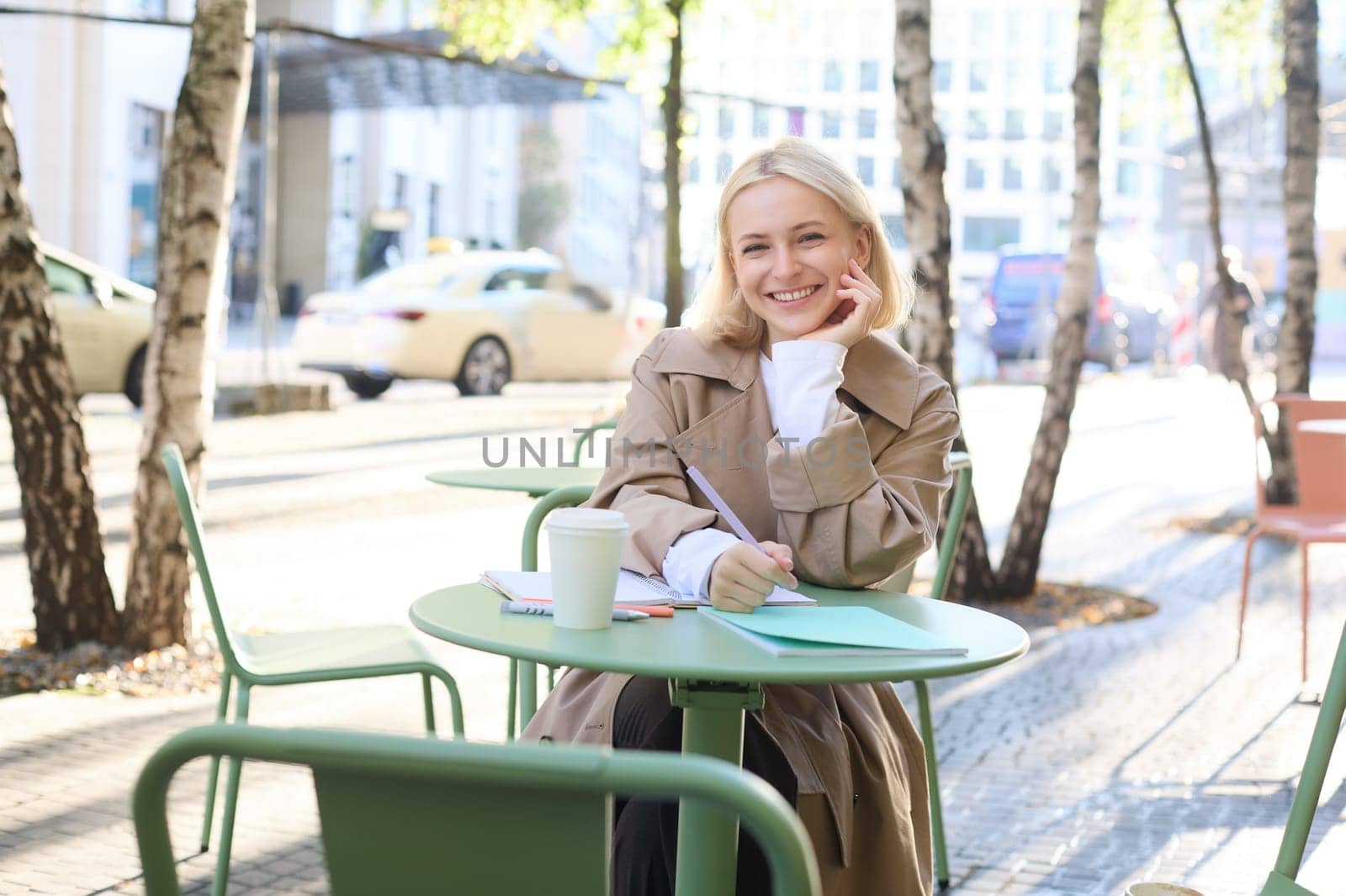 Portrait of young modern woman in stylish trench coat, sitting in outdoor cafe on sunny day, writing in her notebook, working or doing homework, drinking coffee by Benzoix