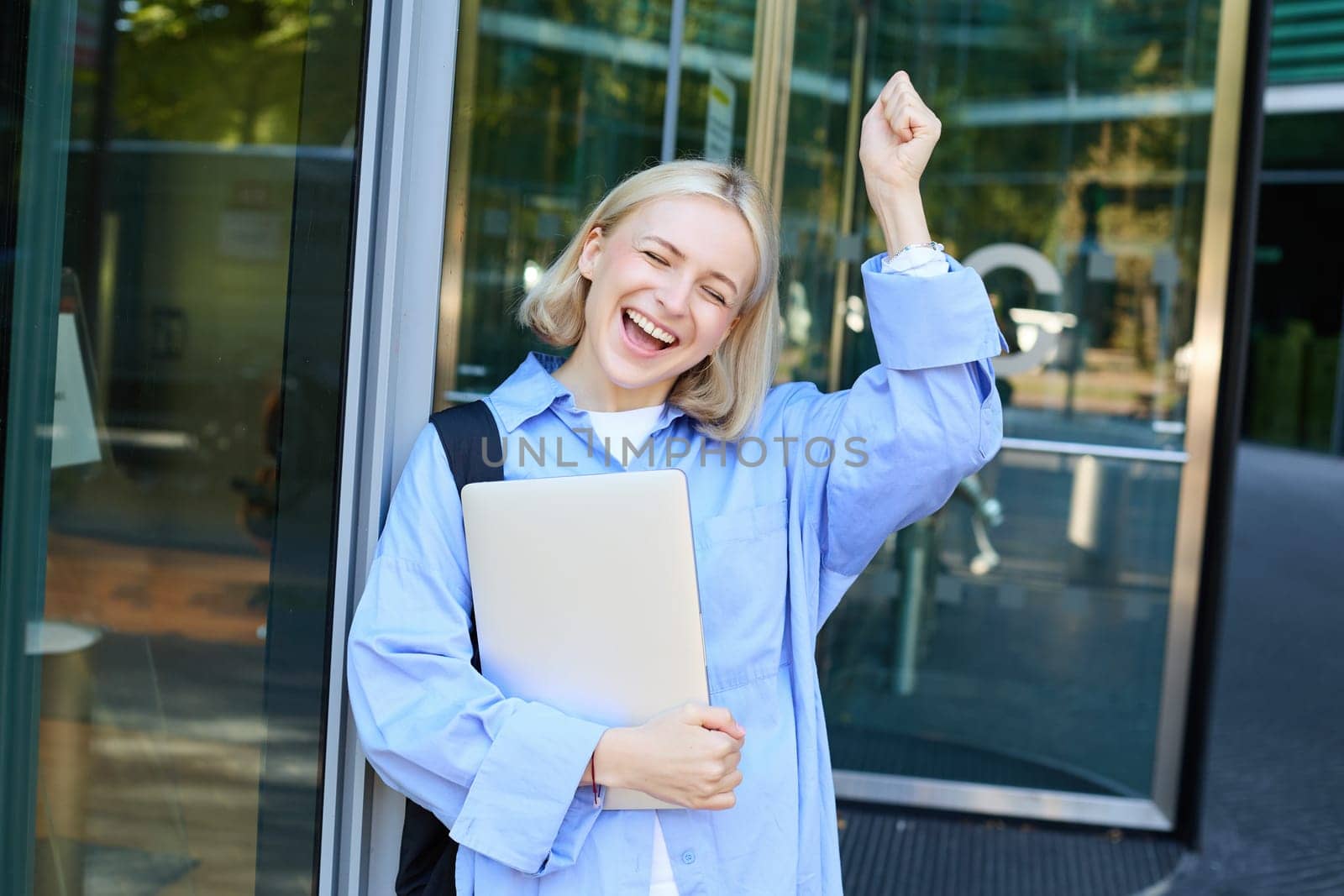 Carefree blond woman laughing, smiling and celebrating, posing with laptop near office, campus building, raising hand up in triumph, feeling excited by Benzoix