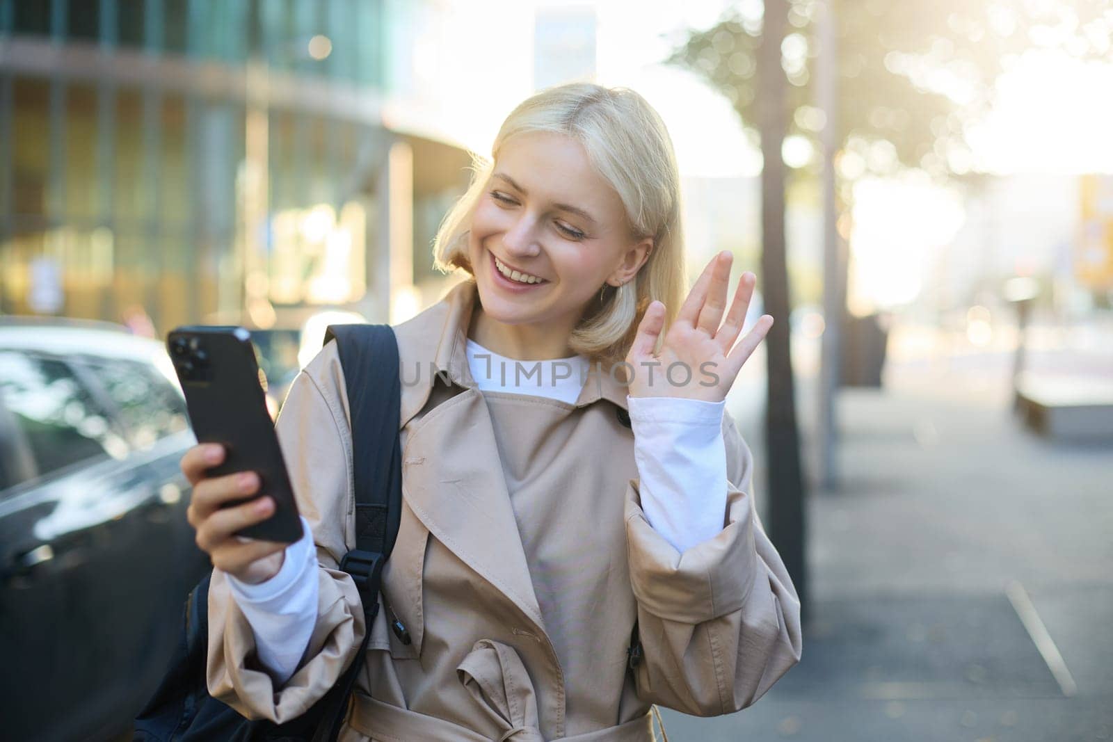 Smiling young modern woman, walking on street, video chats with friend, says hello, waves hand at mobile phone camera, talking to someone on smartphone application.