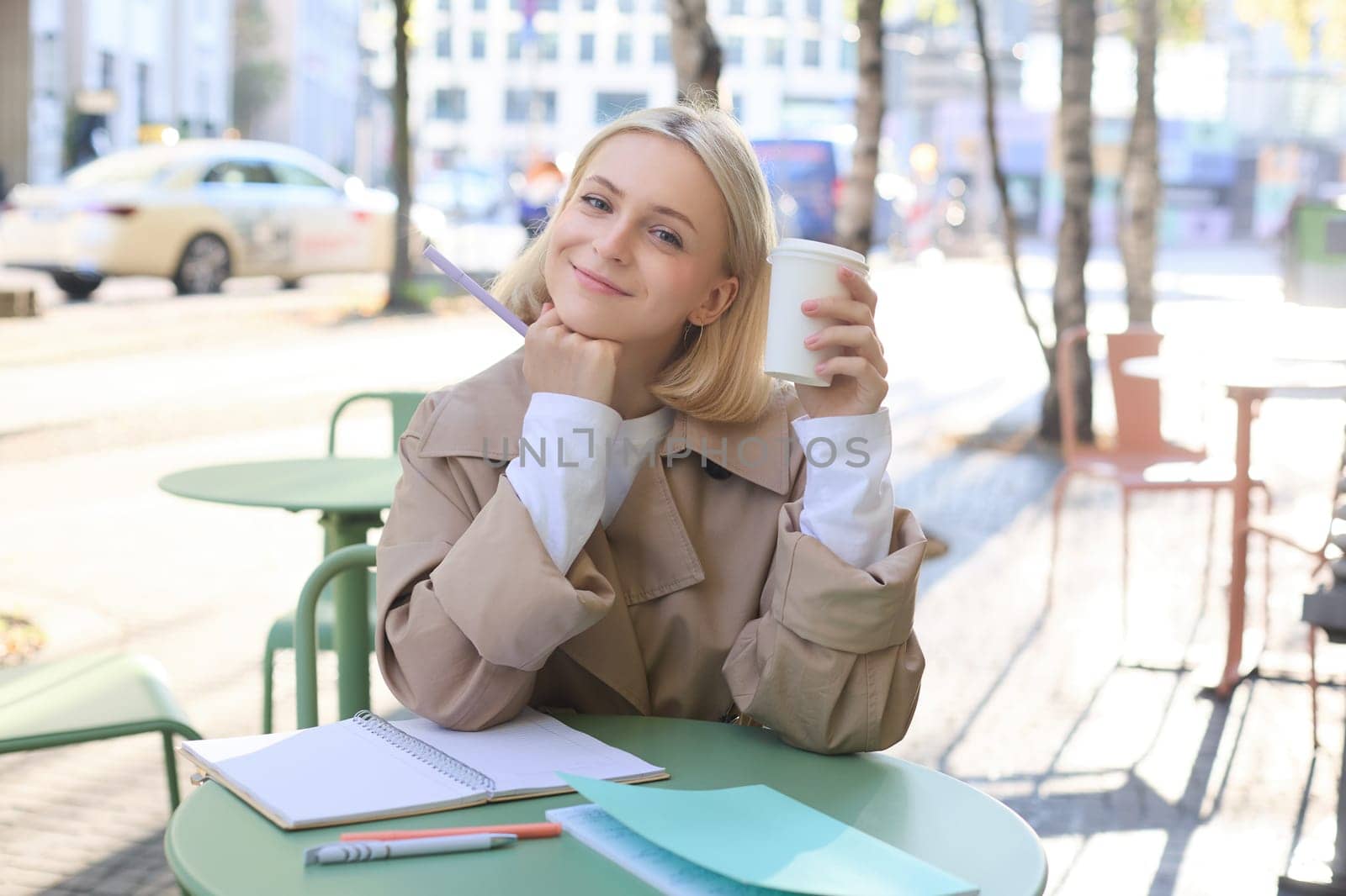 Image of beautiful young blond woman, girl drinking coffee in outdoor cafe, working on university project, doing homework, enjoying sunny weather, smiling at camera.