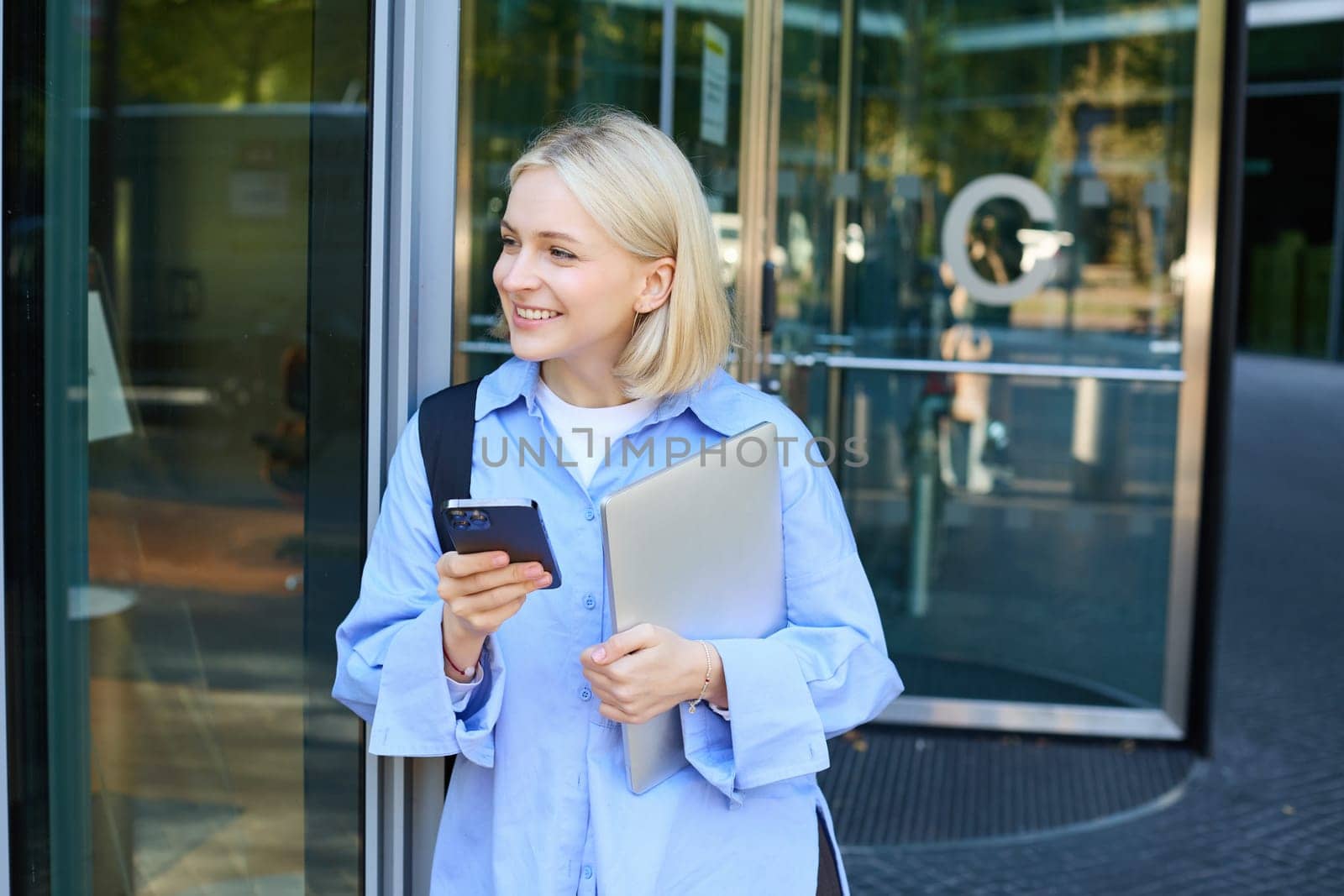 Image of stylish young woman, student walking out of the college campus, holding smartphone and laptop, has backpack on shoulder, waiting for someone outside with mobile phone by Benzoix