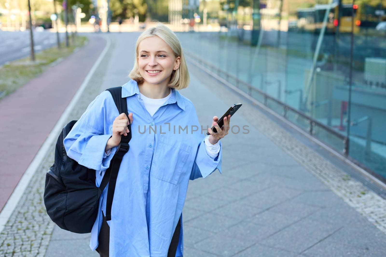 Lifestyle portrait of smiling blond woman with smartphone, walking on street with backpack and looking happy, on her way to college, using mobile phone by Benzoix