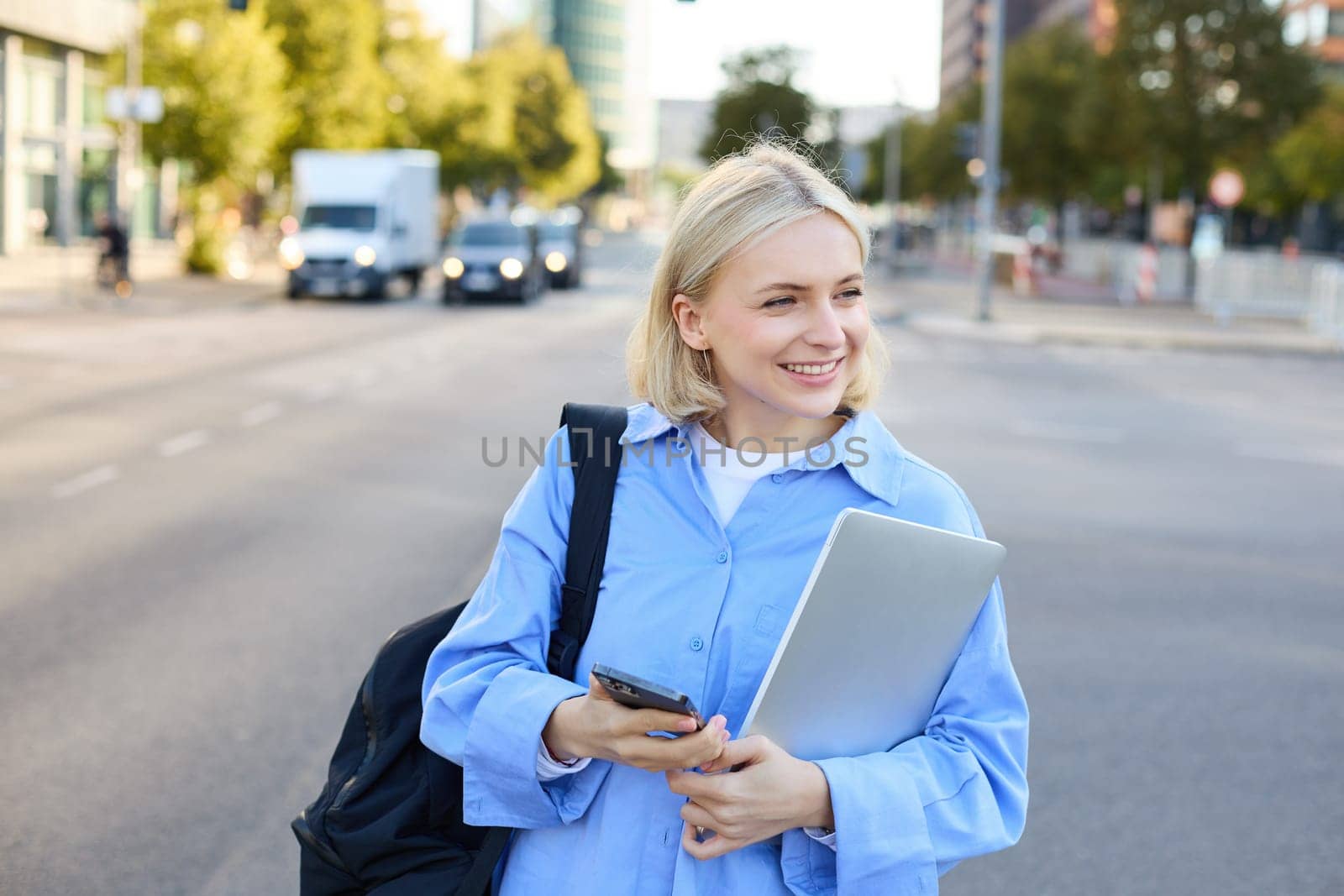 Portrait of blond smiling woman in blue shirt, holding backpack and smartphone, waiting for her ride, order taxi and checking updates on mobile phone, standing on street near road by Benzoix