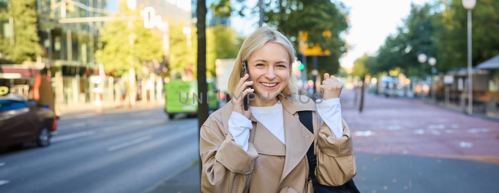 Close up portrait of beautiful young woman, blonge girl walking on street with mobile phone, chatting with friend, has happy face expression while talking over cellphone on her way by Benzoix