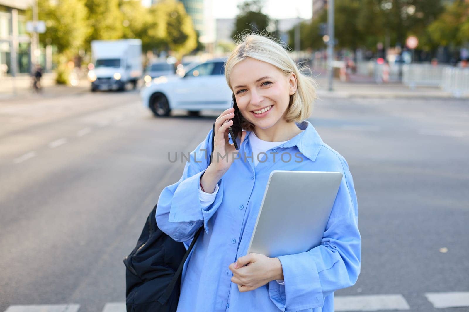 Portrait of young modern female model in blue shirt, talking on mobile phone, chatting with someone while walking along the street, holding laptop. Lifestyle and people concept