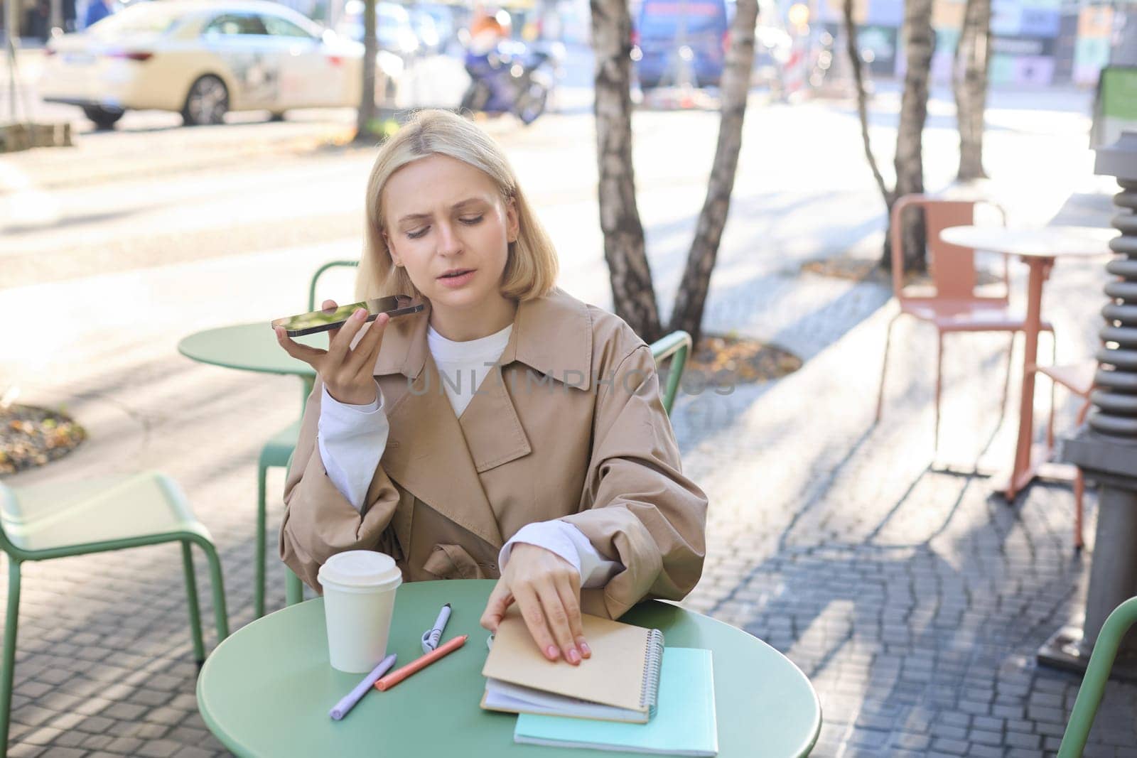 Portrait of serious, concentrated woman, opens her notebook, records voice message, sending notification to coworker, drinking coffee, working outdoors by Benzoix