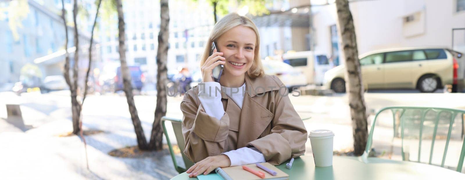 Close up of cheerful blond girl, woman talking on mobile phone, sitting in city centre, outdoor cafe, taking break from work or studying, chatting on telephone.