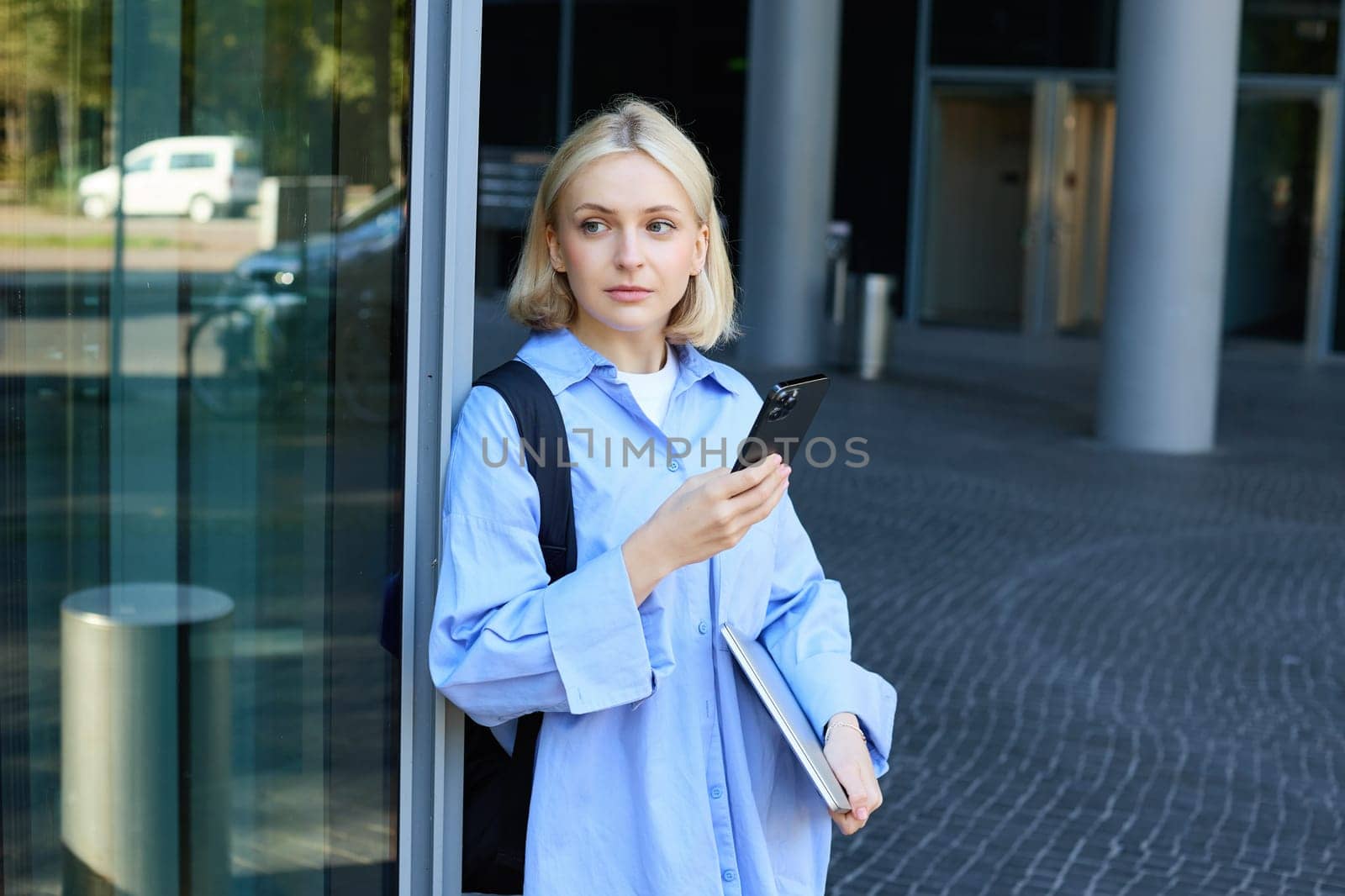 Portrait of young female student standing outside university campus, posing near building, holding laptop and smartphone, wearing blue collar shirt by Benzoix