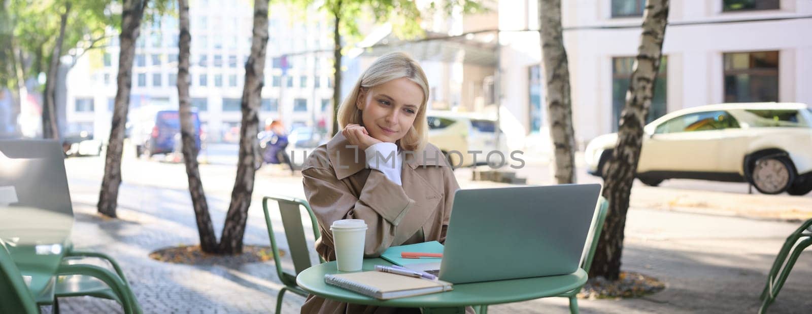 Image of young woman looking at laptop, sitting in outdoor cafe and drinking coffee, studying remotely, attends online course while sits outside, working, freelancing.