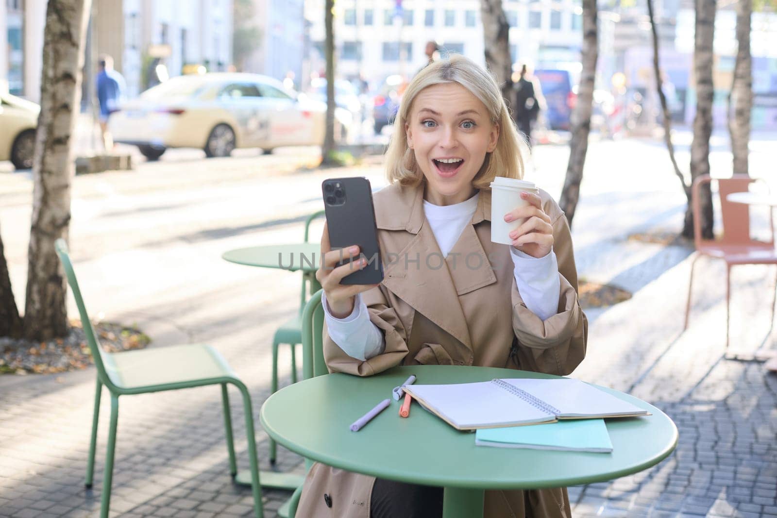 Image of girl with coffee and smartphone, sitting on street, looking surprised, gasping at camera.