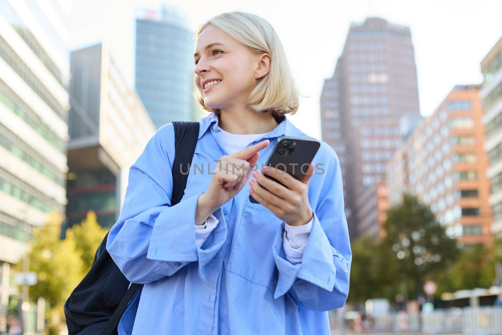 Close up portrait of stylish young blonde woman, standing on street, checking her mobile phone, using smartphone app to get around town, looking at online map for directions by Benzoix