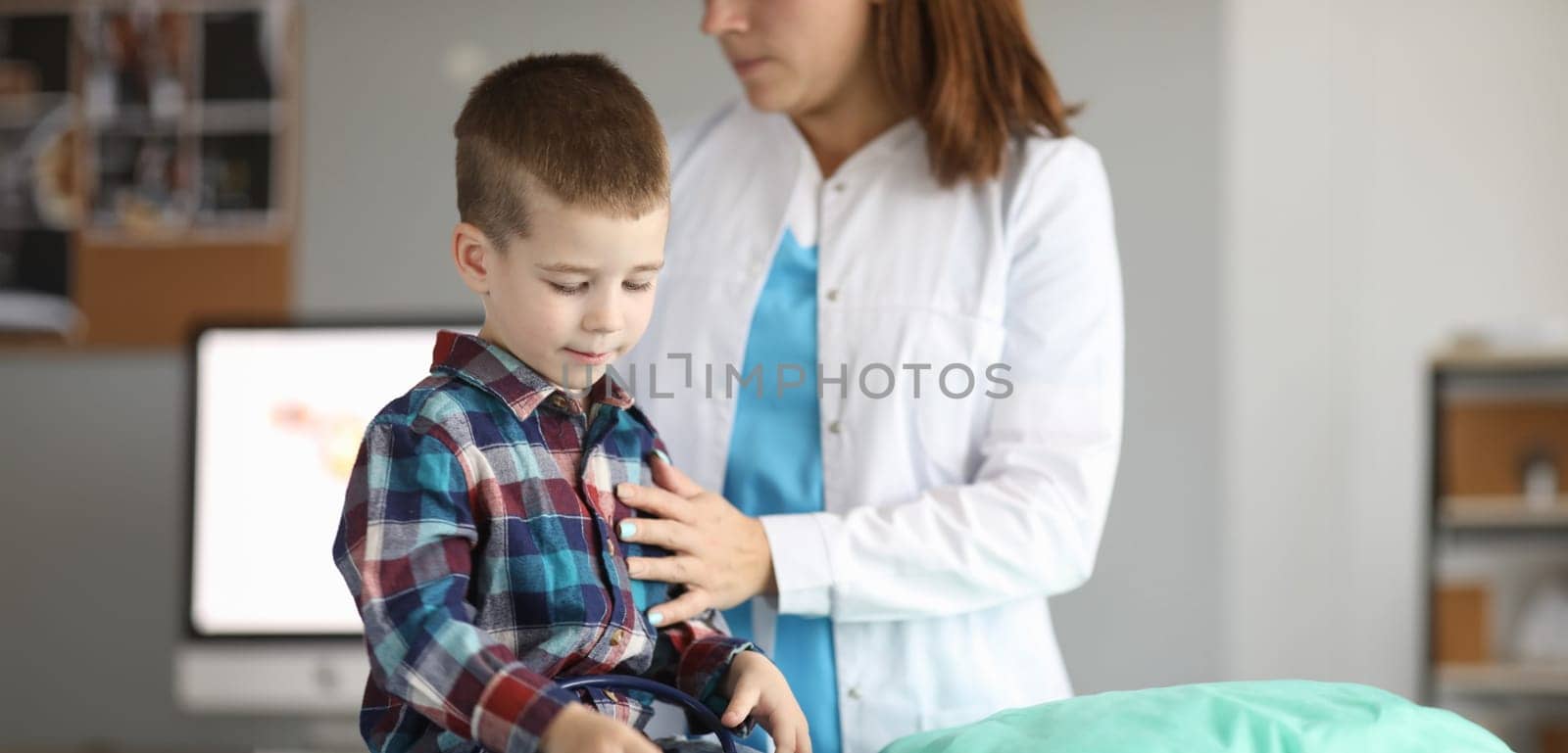 Portrait of child sitting in modern clinic office and looking down in gloomy mood. Female pediatrician standing near patient and examining boy. Medicine and healthcare concept