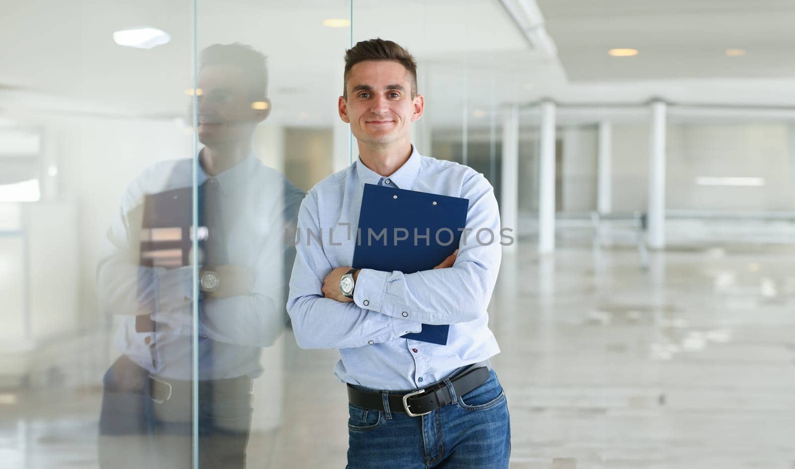 Handsome man in shirt and folder stand in office looking in camera hands crossed on chest. White collar dress codemodern office lifestyle graduate college study profession idea coach train concept