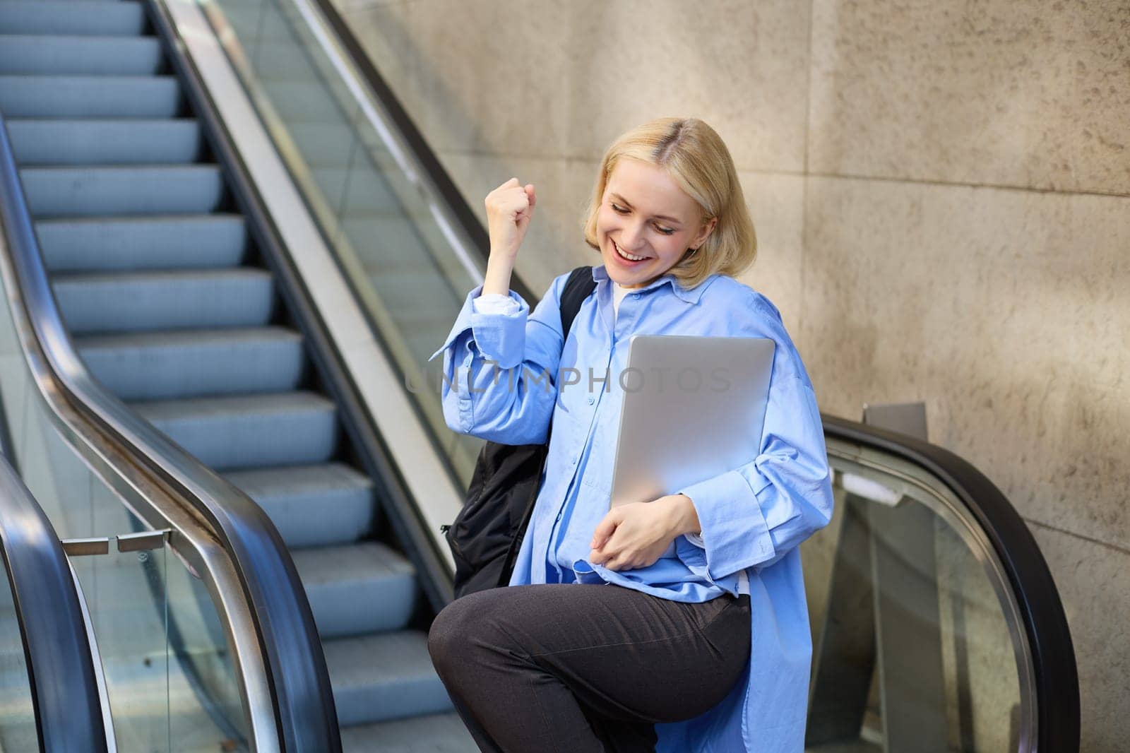 Excited young blond woman with backpack and laptop, says yes, winning, celebrating victory, achieve goal, posing near escalator. Lifestyle concept