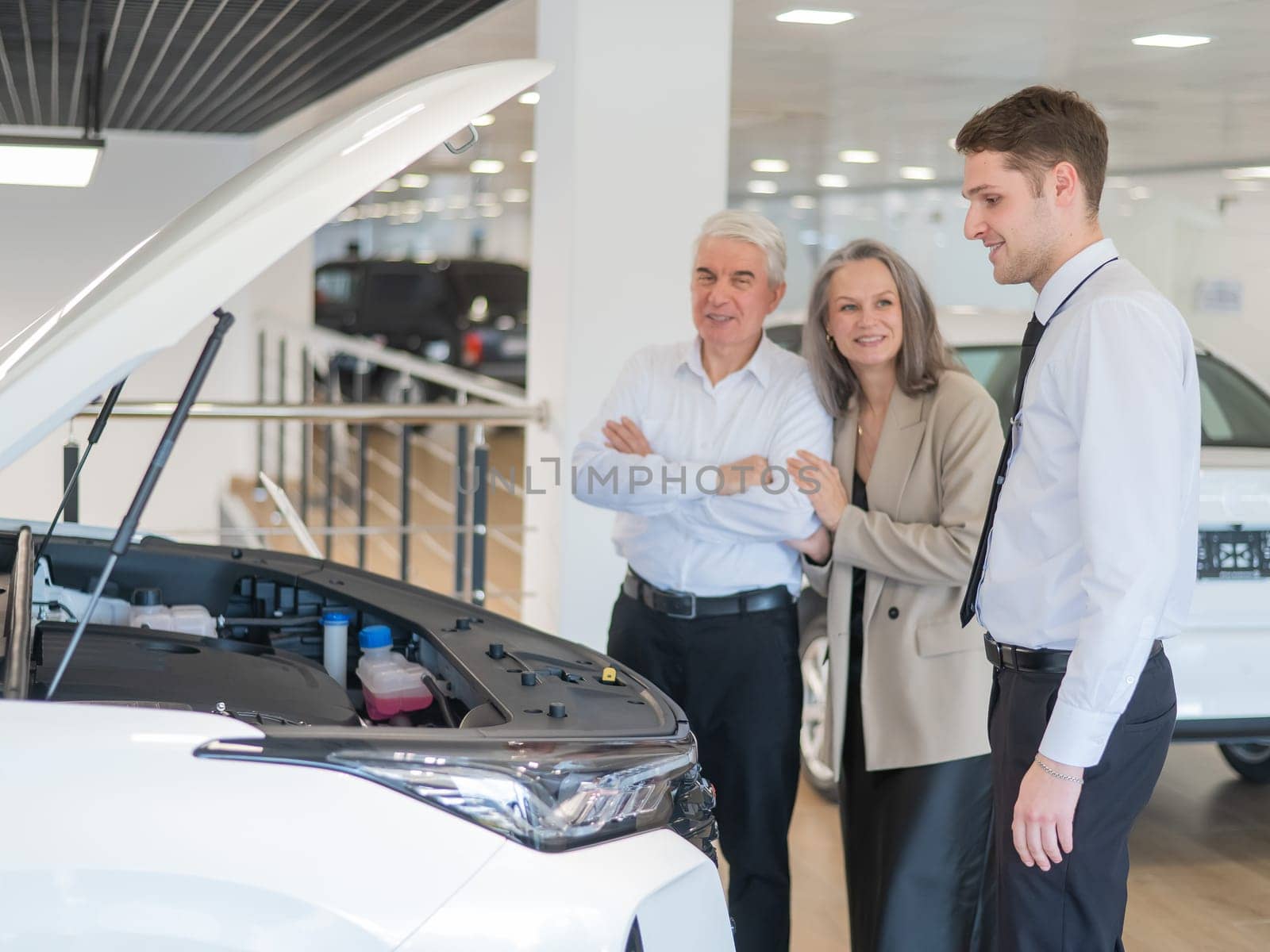 A salesman demonstrates a car with an open hood to an elderly Caucasian couple in a car dealership. by mrwed54