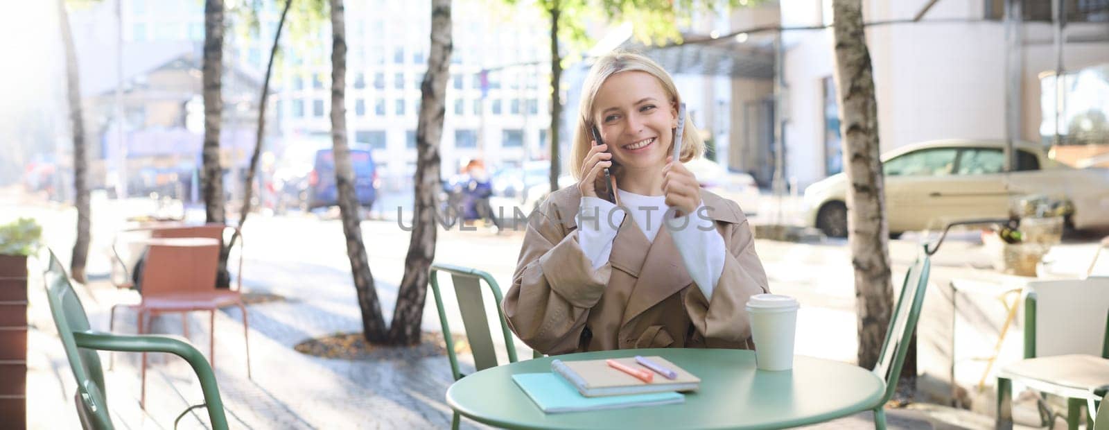 Lifestyle and urban people concept. Young smiling girl, student sitting in cafe outdoors, talking on mobile phone, chatting with friend or coworker over the telephone, studying or doing homework by Benzoix