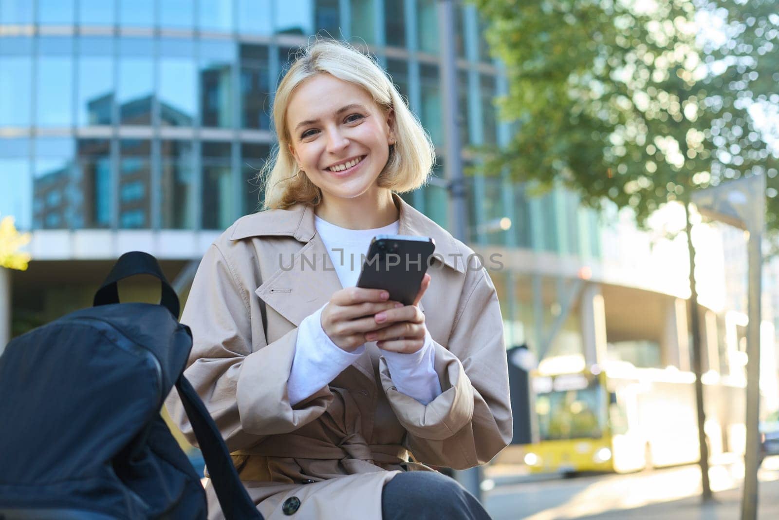 Lifestyle portrait of cute blonde girl with smartphone, sitting on bench with backpack, using mobile phone, reading message.